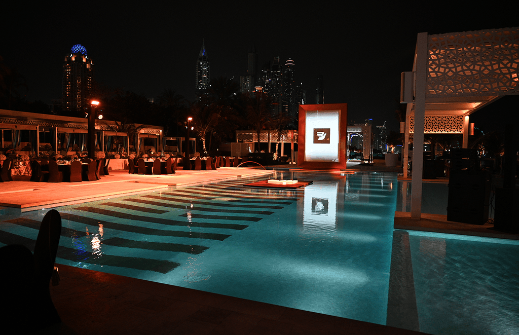 A beautifully lit poolside setting in Dubai, featuring elegant tables arranged for a company event with dinner and drinks, against the backdrop of an illuminated cityscape.