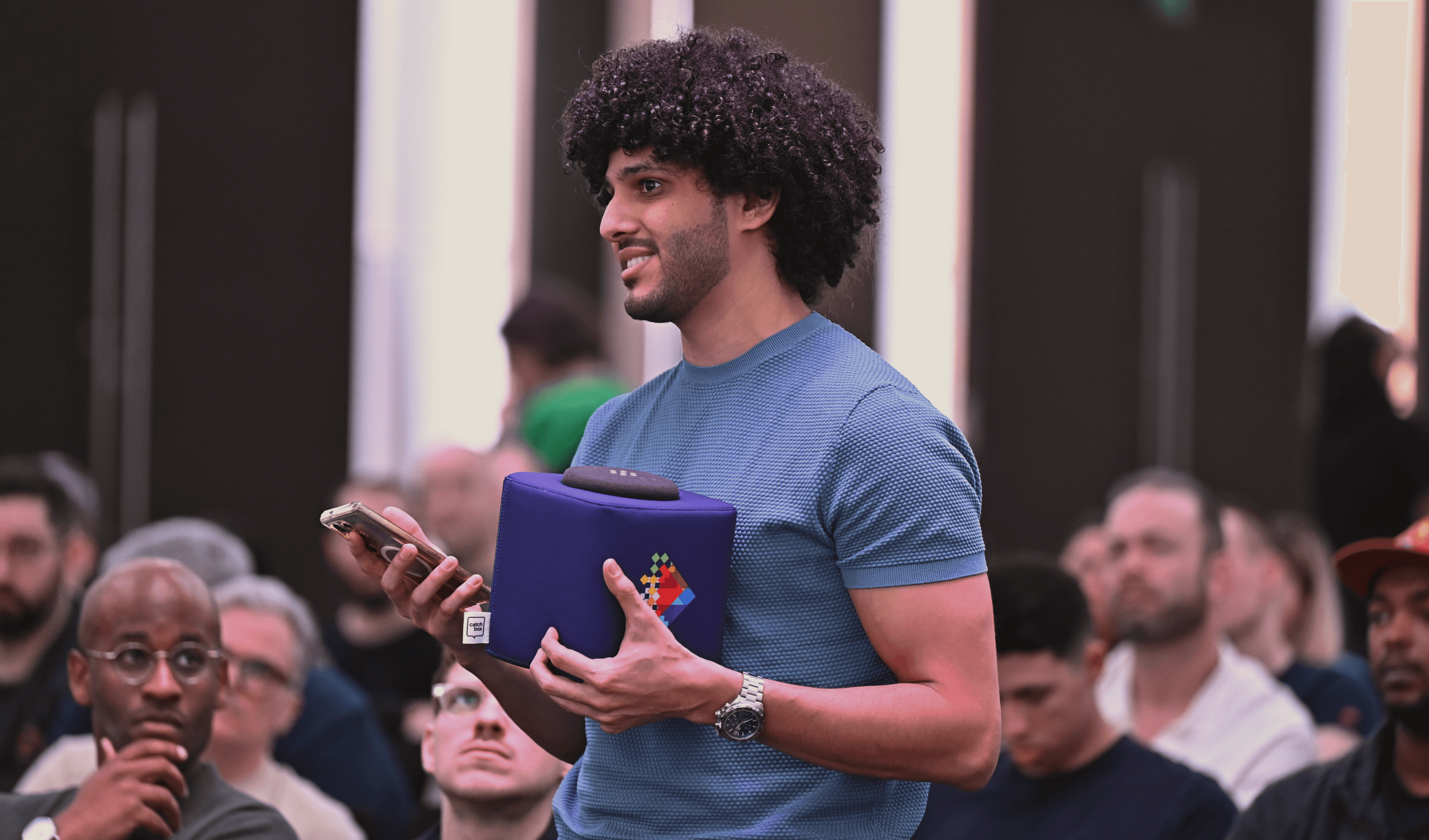 In a bustling Dubai event, a man in a blue shirt actively participates during question time by holding a microphone amidst a diverse audience.