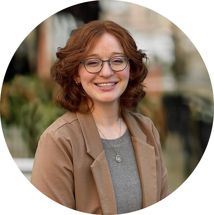 Smiling woman with curly red hair, wearing a brown blazer, stands outdoors with greenery in the background.